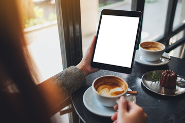 Mockup image of a woman sitting and holding black tablet pc with blank white desktop screen while drinking coffee in cafe