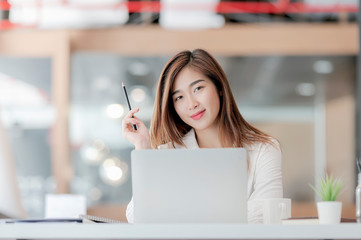 Portrait of young pretty businesswoman holding pencil, smiling and looking at camera while sitting at office desk.