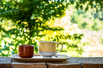 Cup of coffee and tea on wooden plate over blurred plantation of tree with sun lighting.