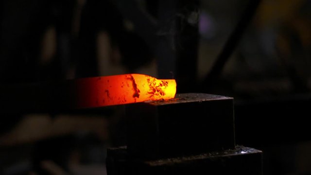 Worker uses forge hammer in metal workshop. Glowing hot steel is slowly shape to form a knife blade, as hammer impacts down on anvil. Metal deforms and flakes away in slow motion.