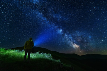 Naklejka na ściany i meble Bright starry sky with the milky way on the background of mountains and hiker with red tent.