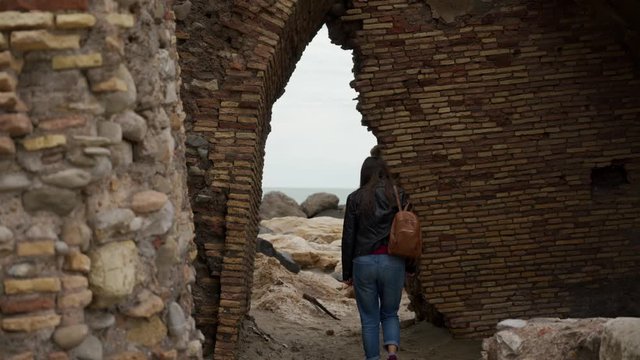 Tourist girl with a backpack goes through arch in ruins of an ancient fortress Torre Flavia in Ladispoli, Italy. Rear view