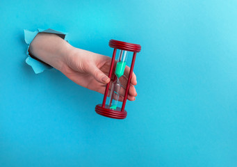 Hourglass in a female hand on a blue background with a hole, time concept