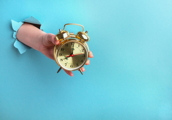 Alarm clock in a female hand on a blue background with a hole, time concept