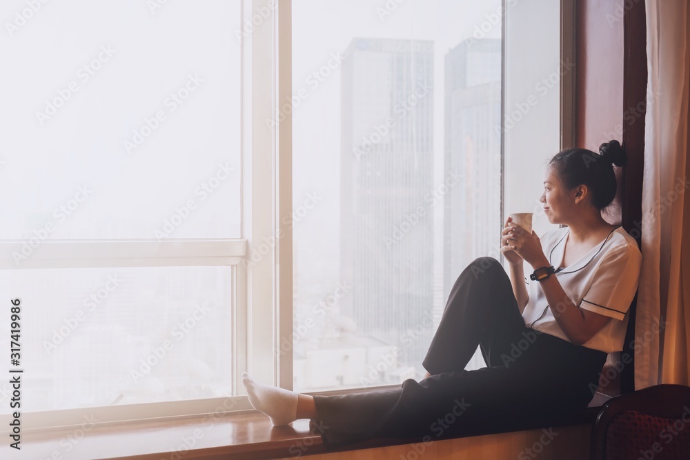 Wall mural asian beautiful woman thinking while drink coffee and sitting in room with building outside window b