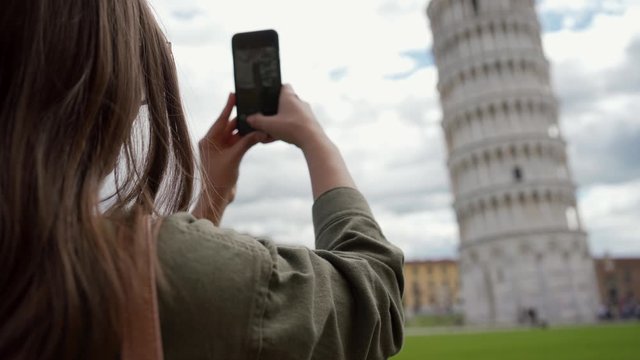 Young female tourist taking vertical pictures of the famous Leaning Tower of Pisa against cloudy sky using mobile phone. Rear view. Vacation in Italy, traveling season.