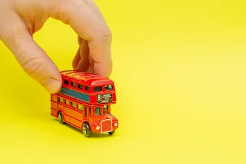 Fototapeten Britischer Spielzeug-Doppeldecker-roter Bus, der von männlicher Hand auf gelbem Hintergrund fährt. Konzept des Englischunterrichts und Verbesserung der Sprech- und Sprechfähigkeiten © dddoria