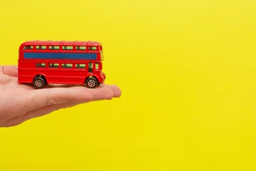 Fototapeten Britischer roter Spielzeug-Doppeldeckerbus, der von männlicher Hand auf gelbem Hintergrund mit Kopienraum für Ihren Text hält. Konzept des Englischunterrichts © dddoria