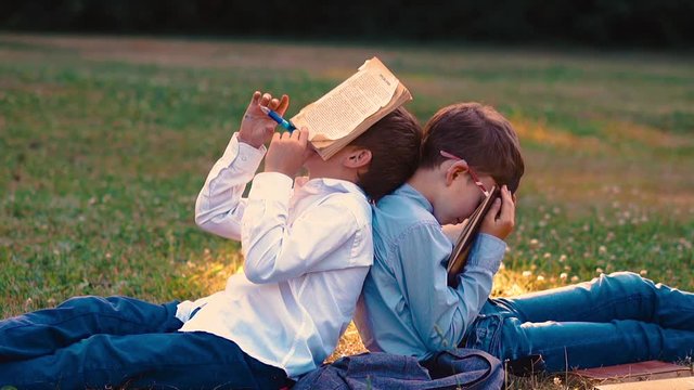 sleeping student fixes pen in mouth to hold book leaning on schoolmate dreaming with book on green garden lawn slow motion