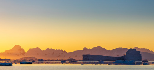 Panorama of golden yellow sunset over the mountains, water, and icebergs of Antarctica, a pristine remote landscape © Gabi