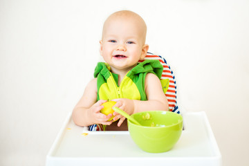 Little happy baby with spoon sits at highchair and eats porridge on plate