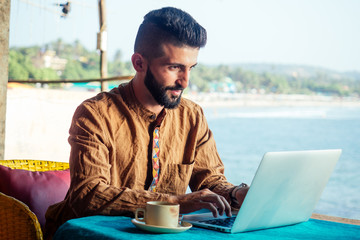 Egyptian happy man with laptop computer drinking coffee or masala chai at beach shack in Goa