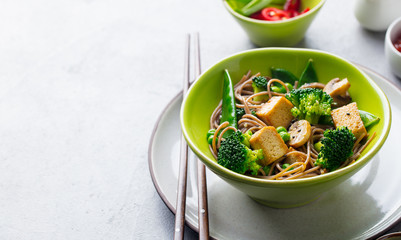 Soba noodles with vegetables and fried tofu in a bowl. Copy space. Grey background.