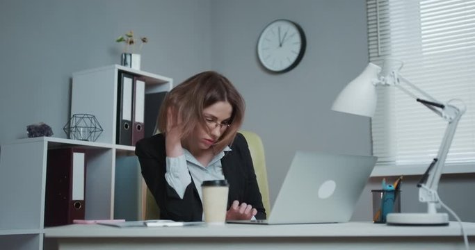 Tired woman in glasses sitting at the laptop computer while working in the office, then almost falling asleep and waking up