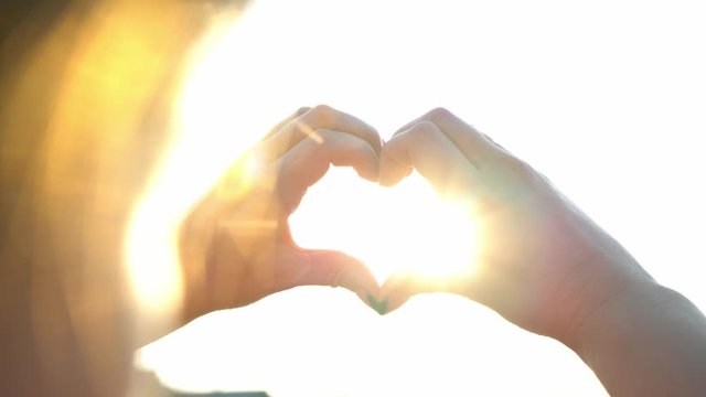 Woman makes hands in the shape of a heart in the bright sun. The magical rays of the sun shine through your fingers. Fantastic bright light. Symbol of love. Close up of attractive woman hands.