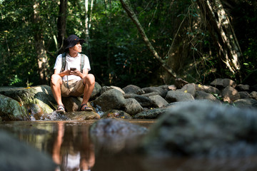 Fototapeta na wymiar A male hiker sitting on rock near stream using mobile phone