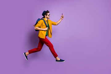 Fototapeta na wymiar Full size profile side photo of cheerful guy in retro headwear holding blue bag jump run make selfie on smartphone wear yellow shirt red pants trousers isolated over violet color background