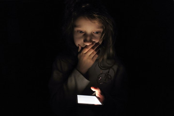 Child girl with a phone in the dark. Frightened child looks at the screen of a smartphone.