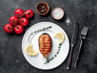 Grilled salmon steak in a white plate with spices, fork and knife on a stone background