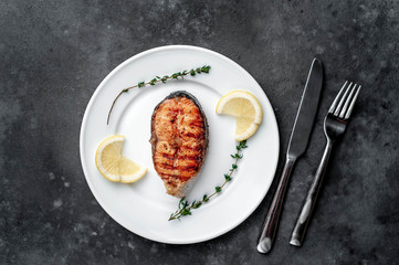 Grilled salmon steak in a white plate with spices, fork and knife on a stone background