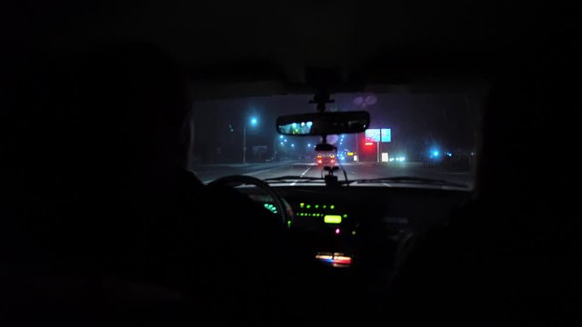 The driver rides at night on a lighted road. The car cracks and staggers. Inside view. Dashboard, radio, DVR. Overtaking. Oncoming cars blind eyes. Headlights light the way. Road signs and marking.