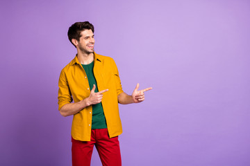 Photo of toothy beaming cheerful man pointing into empty space to show you necessary information in red trousers isolated violet pastel color background