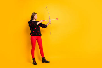 Full length profile photo of pretty lady working as cupid hold bow arrow aiming feelings love couple wear hearts pattern sweater red trousers shoes isolated yellow color background