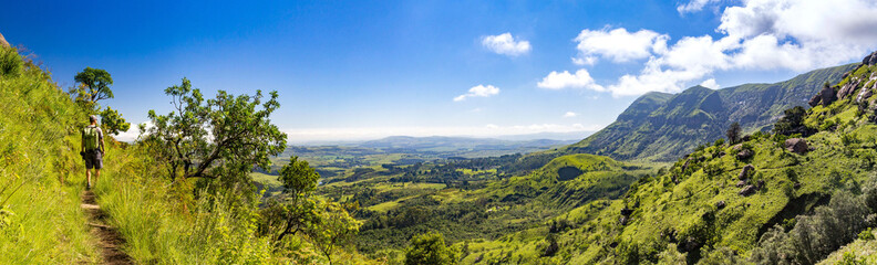 Man hiking on a small trail with a panoramic view over steep and green mountains on a sunny day,...
