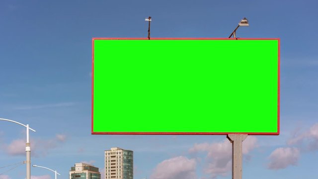 green chroma-key billboard against modern building tops under blue sky with clouds on sunny day zoom in