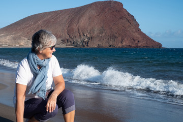Fototapeta na wymiar Side view of one caucasian senior female enjoying the beach with the wind in a winter day. Blue sea and red mountain in the background