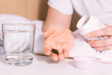 Unrecognizable woman sitting on bed and holding antiviral pills for flu before intake. Glass of...