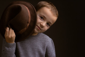 Studio portrait of boy in a gray sweater, on a black background, holding a brown hat behind the brim, hiding and peeking out from behind it