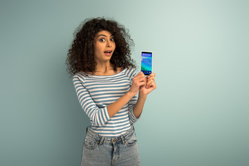 surprised mixed race girl showing smartphone with booking app on grey background