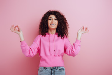 cheerful bi-racial girl looking at camera while standing in meditation pose on pink background