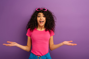 disappointed bi-racial girl showing shrug gesture while looking at camera on purple background