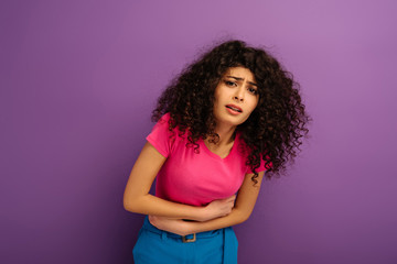 upset bi-racial girl touching stomach while suffering from abdominal pain on purple background