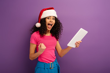 excited mixed race girl in santa hat showing winner gesture while holding digital tablet on purple background