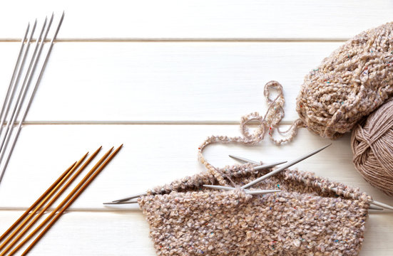 Circular knitting for a warm "bini" hat with five knit needles from boucle wool yarn and sets of knitting needles on a white wooden table. Copy space, flat lay, top view