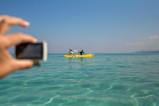 Father And Son Having Fun During Summer Beach Vacations. Blurry Female Hand Of Mother Holding Action Camera Shooting Video Of Family Adventures. Boy And Man Kayaking Together In Sunny Sea Water.