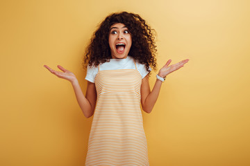 shocked mixed race girl looking away while standing with open arms on yellow background