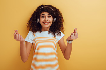 excited mixed race girl in wireless headphones showing winner gesture on yellow background