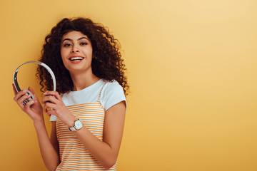 happy bi-racial girl smiling at camera while holding wireless headphones on yellow background