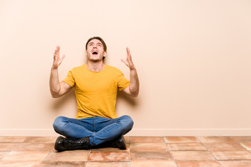 Young caucasian man sitting on the floor isolated screaming to the sky, looking up, frustrated.