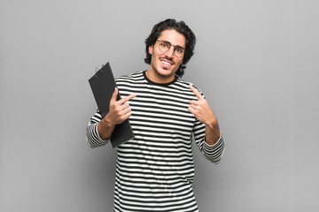 Young employee man holding an inventory smiles, pointing fingers at mouth.