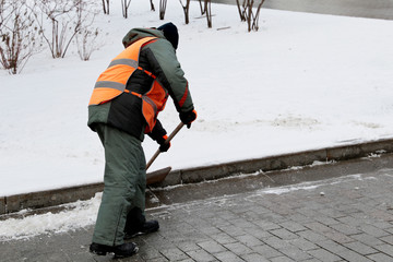 Snow removal in winter, communal services worker in uniform with a shovel clears snow on a street