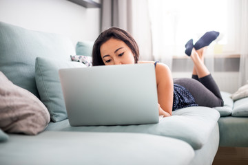 Smiling asian young woman using laptop lying on sofa at home