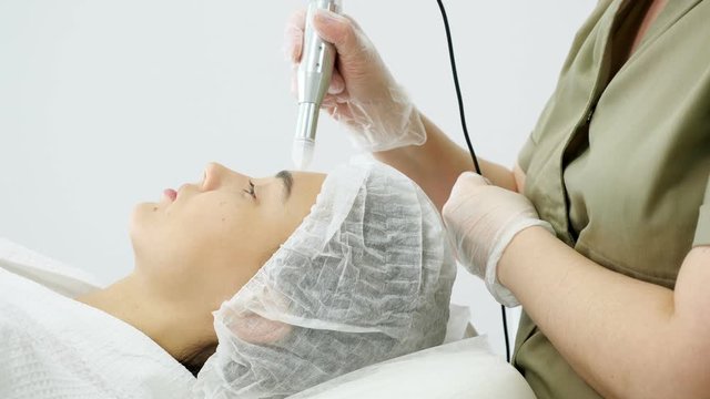 beautician hand moves microdermabrasion device on young woman forehead against white clinic wall close-up