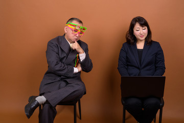 Obraz premium Funny mature Asian businessman with mature Asian businesswoman using laptop