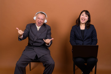 Obraz premium Mature Asian businessman listening to music with mature Asian businesswoman using laptop