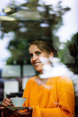 Young woman laughingly drinking tea or coffee in a cafe behind a glass pane
